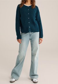 Tyrkysový pletený cardigan s dlouhými rukávy a kulatým výstřihem, zdobený bílými knoflíky. Kombinován s světle modrými širokými džínami.