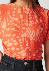 Blusa de manga corta en tejido texturizado naranja con un patrón floral en crema. Cuenta con un cuello redondo y un detalle de botón en la parte delantera.
