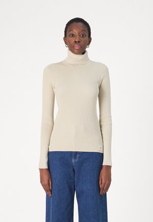 Beige ribbed turtleneck-paita pitkillä hihoilla, jossa on istuva malli ja huomaamaton brändäys alareunassa, yhdistettynä leveälahkeisiin farkkuihin.