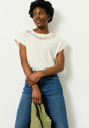 Weißes kurzärmeliges Oberteil mit floraler Stickerei am Ausschnitt, kombiniert mit hochtaillierten blauen Jeans. Accessoire: olivgrüne Jacke.