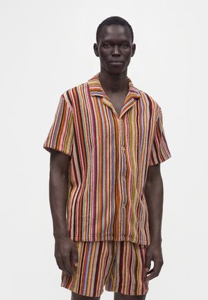 Hombre con camisa de manga corta abotonada y pantalones cortos a juego con rayas verticales en tonos rojos, amarillos, azules y marrones.