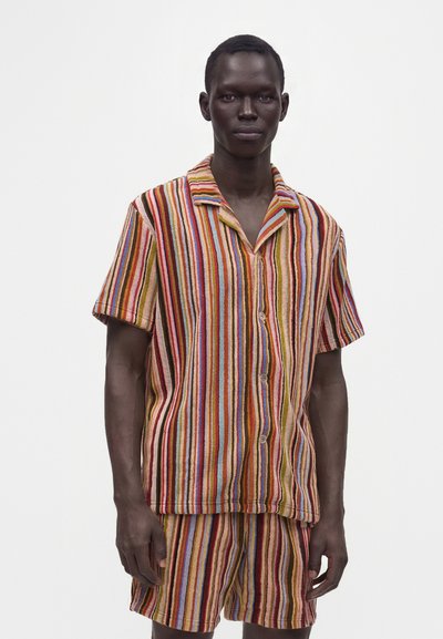 Homme portant une chemise à manches courtes boutonnée et un short assorti à rayures verticales rouges, jaunes, bleues et marron.