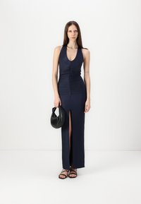 Marine sateng halterneck kjole med rynkede detaljer og sidesliss, kombinert med en liten svart veske og tynne svarte sandaler.