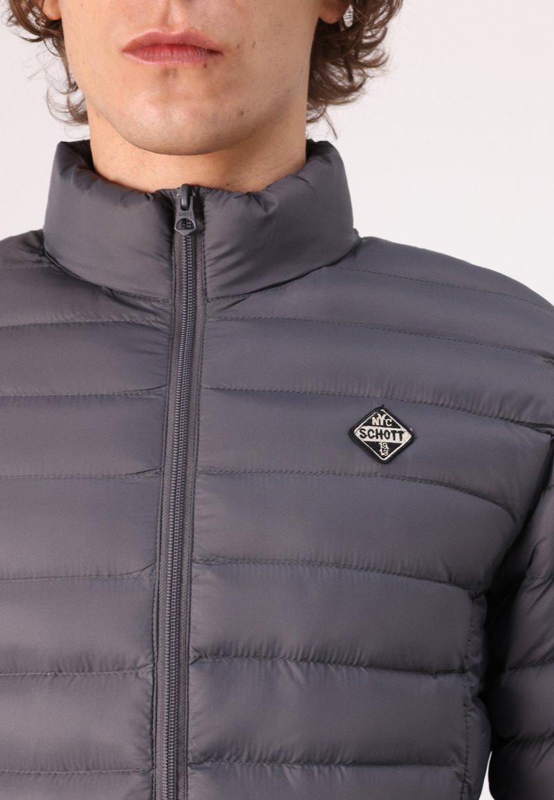 Doudoune grise avec col haut, fermeture éclair et coutures horizontales. Présente un patch logo en noir et blanc sur la poitrine.