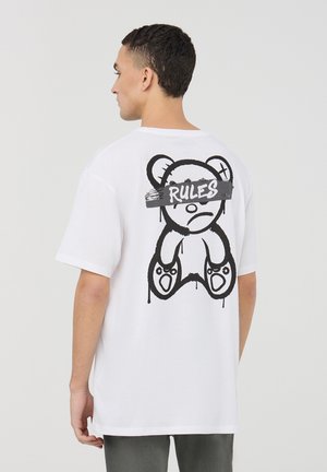 T-shirt bianco in cotone con una grande grafica di orso nero sul retro, con la parola "REGOLI" scritta in uno stile di pennellata sopra gli occhi.