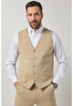 Hombre con barba que lleva un chaleco beige abotonado sobre una camisa blanca, pantalones beige y un reloj, de pie frente a un fondo claro y liso.