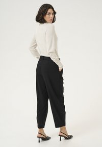 Hellbeige langärmliges Oberteil mit einem subtilen Glanz, kombiniert mit schwarzen maßgeschneiderten Hosen mit seitlichen Taschen und schwarzen Plateausandalen.
