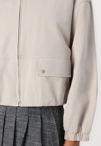 Lys beige cropped jakke med lynlås, elastiske manchetter og en trykknap-lomme detalje. Lavet af et glat, letvægtsmateriale.
