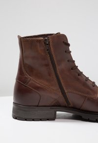 Bottine en cuir marron avec une fermeture éclair sur le côté, présentant un avant lacé, une surface texturée et une semelle en caoutchouc robuste avec un motif de relief.