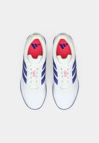 Zapatos deportivos blancos con tres franjas moradas, una parte superior sintética texturizada y una plantilla roja vibrante con un logo.