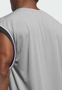 Top deportivo gris de tirantes con una tela de malla texturizada, que presenta acentos en blanco y negro en las sisas y en el cuello, mostrando la vista trasera.