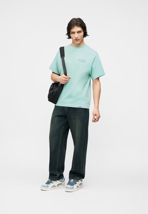 Nuori mies pukeutuneena mintunvihreään, ylisuurikokoiseen T-paitaan, tummiin väljälahkeisiin farkkuihin, valkoisiin ja sinisiin lenkkareihin sekä mustaan olkalaukkuun, seisomassa yksiväristä taustaa vasten.