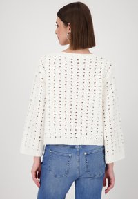 Crocheted white sweater with a loose fit, featuring open knit patterns, wide sleeves, and a ribbed hem, paired with blue denim jeans.