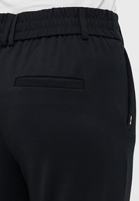 Pantalons de survêtement noirs en tissu doux et extensible, dotés d'une taille élastique, d'une poche plaquée arrière et d'un petit accent métallique sur le côté.