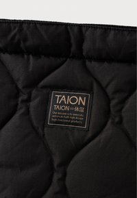 Μαύρο πικέ ύφασμα με υφή και εξέχουσα ορθογώνια ετικέτα. Η ετικέτα διαθέτει χρυσό κείμενο που αναφέρει την επωνυμία και την αποστολή.