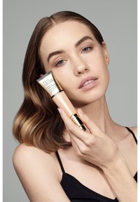 Beige tube van Estée Lauder product met gouden accenten, vastgehouden tegen een neutrale achtergrond. Model heeft middelhoofbruin haar en een natuurlijke make-uplook.