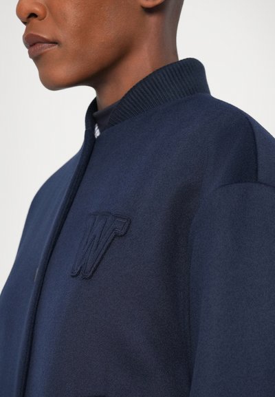 Námornícky vlnový bomber s rebrovým golierom, s vyšitým logom "W". Hladká textúra s štruktúrovaným dizajnom.