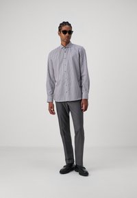 Gekookte button-up blouse in wit en marineblauw, gecombineerd met slanke grijze broek. Model draagt zwarte zonnebril en zwarte schoenen.