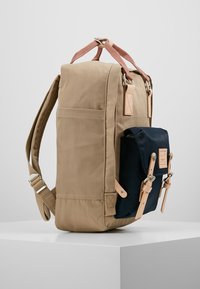 Beżowy plecak o wyraźnym kształcie, z skórzanymi akcentami i ciemnoniebieską przednią kieszenią. Posiada wyściełane szelki oraz detale po bokach.