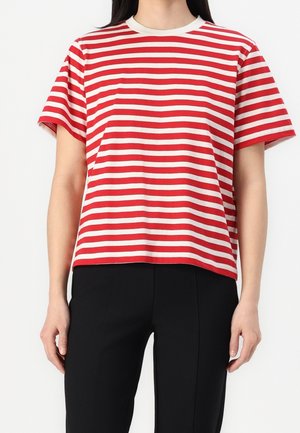 Person iført en rød og hvid vandret striped kortærmet skjorte og sorte bukser, stående mod en ensartet hvid baggrund.