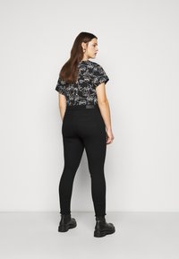 Camiseta negra con patrones abstractos de caras en blanco, combinada con jeans ajustados negros y botas de tobillo negras, vista desde la espalda.