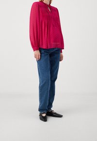 Blouse fuchsia à manches longues avec plissé et col à nouer, associée à un jean bleu droit et des ballerines noires pointues. Motif à pois sur la blouse.