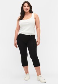 Zwarte capri leggings gecombineerd met een witte, mouwloze top. Het materiaal lijkt zacht en rekbaar. Het model draagt witte sneakers. Eenvoudig ontwerp, zonder patronen.