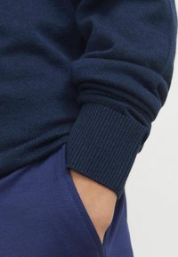 Manche de pull en tricot bleu marine avec poignet côtelé, couvrant partiellement une main dans une poche violette de vêtements assortis. Texture douce évidente.