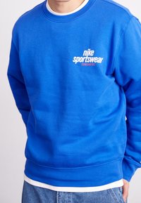 Blå tröja i bomullsmix med ribbade ärmslut och nederkant. Har en vit Nike Sportswear-logotyp på bröstet med röda detaljer.