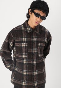 Veste en polaire à carreaux bruns avec un devant boutonné, un col et une poche poitrine, dotée d'une surface texturée et douce. Lunettes de soleil noires portées.