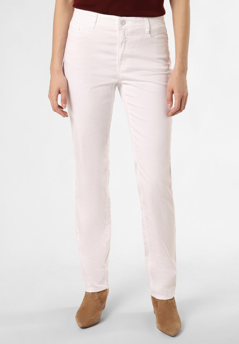 Femme portant un pantalon blanc taille haute et des bottines marron, debout, les mains détendues le long du corps, devant un fond uni.