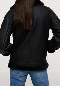 Veste en cuir noir avec un doux bord en fourrure au bas, présentant un design épuré, des manches longues et des détails de couture visibles au dos.