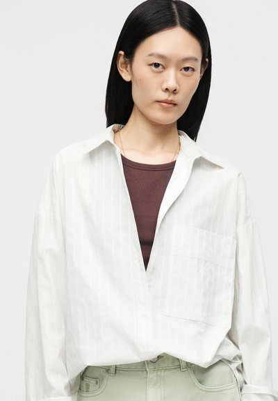 Jeune femme aux cheveux noirs et raides portant une chemise blanche oversize boutonnée sur un haut marron et un pantalon gris clair.