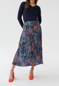 Gonna a pieghe floreale in blu scuro, con tacchi arancioni brillanti. Il materiale ha una texture liscia con motivi floreali colorati su tutto il capo.