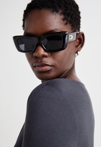 Black oversized sunglasses with a thick frame and dark lenses; gold accent detail on the temples; model wearing a fitted gray top.