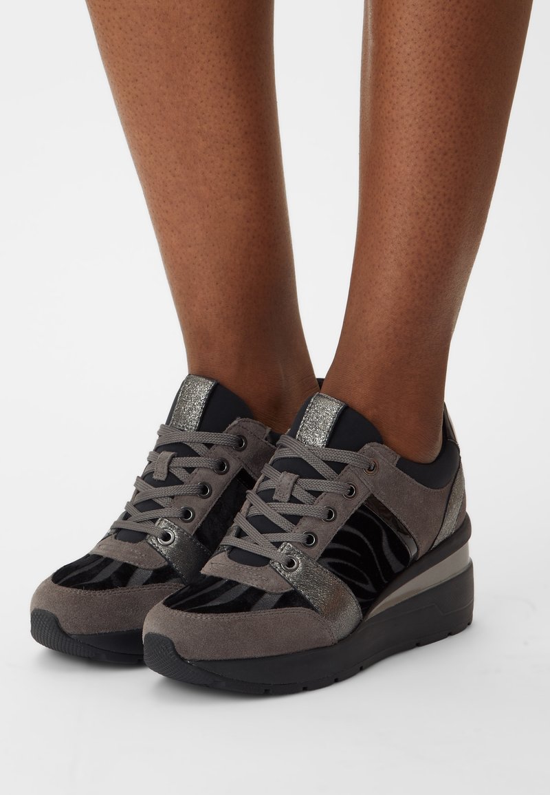 Fötter i svarta och grå plattformsneakers med snörning framtill och metalliska accenter, synliga från mitt på vaden till tårna på en vit bakgrund.