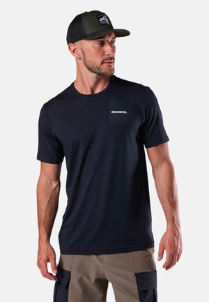 Uomo che indossa una t-shirt nera Rossignol, un cappellino oliva e nero e pantaloncini cargo beige, che guarda verso sinistra su uno sfondo semplice.
