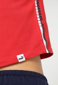 Ourlet de chemise Puma rouge avec bande latérale et étiquette logo au-dessus d'une ceinture élastique bleu marine sur peau nue.