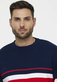 Pull en maille bleu marine avec des rayures horizontales rouges, blanches et rouges. Encolure ronde et tissu texturé. Vue en gros plan de l'avant.