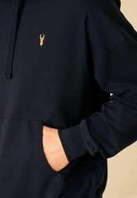 Granatowy hoodie wykonany z miękkiego materiału, z kieszenią kangurzą i małym złotym logo jelonka na klatce piersiowej. Widoczne ściągacze na mankietach i kapturek.