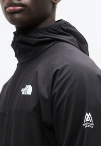 Μαύρο μπουφάν με φερμουάρ, με ψηλό γιακά, λεία υφή και λογότυπα σε λευκό στο στήθος και στο μανίκι. Ισχυρός, αθλητικός σχεδιασμός.