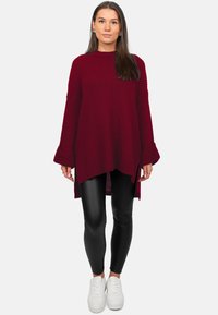 Pull en maille bordeaux à coupe ample, avec texture côtelée, manches longues et ourlet asymétrique, associé à des leggings noirs et des baskets blanches.