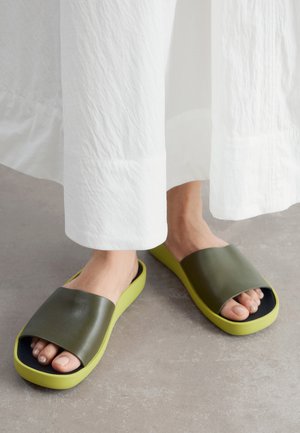 Pieds portant des sandales slides vert olive et jaune avec larges brides, assorties à un pantalon blanc texturé à jambes larges sur un sol gris.