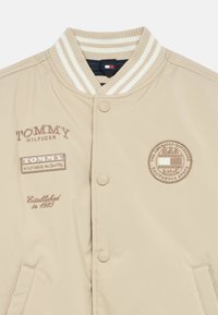 Giubbotto bomber beige chiaro con colletto a coste a righe, dotato di loghi ricamati, bottoni a pressione e tasche laterali.