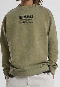 Oliwkowa bluza z haftowanym czarnym napisem „KANI THE ORIGINAL EST. 1989 REG. TRADEMARK” noszona z białymi spodniami i rękami w kieszeniach.