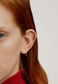 Pendiente de aro dorado con un diseño retorcido, que presenta una superficie texturizada. Llevado en la oreja de un modelo, destacando su estilo minimalista.