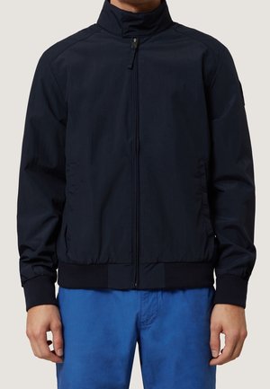 Mann trägt eine schwarze Reißverschlussjacke mit hohem Kragen und gerippten Bündchen, kombiniert mit blauer Hose, gezeigt vom Hals bis zur Hüfte.