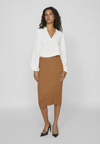 Witte blouse met een V-hals en pofmouwen, gecombineerd met een geribbelde, knielange bruine rok. Zwarte hoge hakken met gouden accenten.