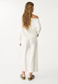 Bela off-shoulder pulover z dolgimi rokavi, v kombinaciji z belimi širokimi hlačami in sproščenimi rjavimi in kremnimi supergami. Teksturovana tkanina. Zadnji pogled.