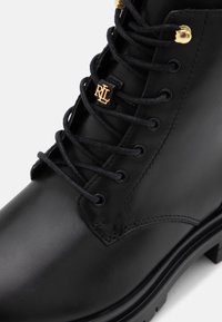 Black leather ankle boot featuring black laces, gold hardware accents, and a textured finish. Includes a logo detail on the front.